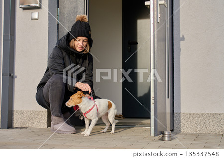 Woman crouching at house entrance putting leash on small dog 135337548