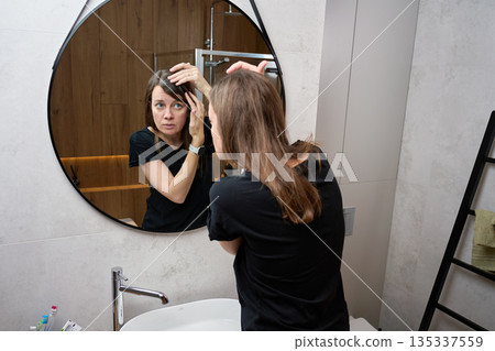 Woman looking at hairline and touching hair while standing in front of bathroom mirror. Woman looking at hairline and touching hair while standing in front of bathroom mirror. 135337559