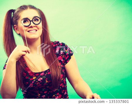 Happy teenage woman holding fake eyeglasses on stick Happy teenage woman holding fake eyeglasses on stick 135337801
