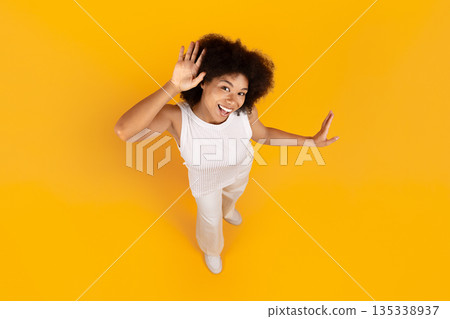 Playful black woman waving and smiling, lifestyle portrait on yellow studio background 135338937