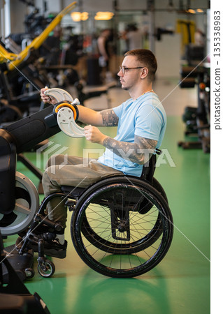 Confident disabled man in wheelchair visiting gym for rehabilitation session after spinal injury. 135338983