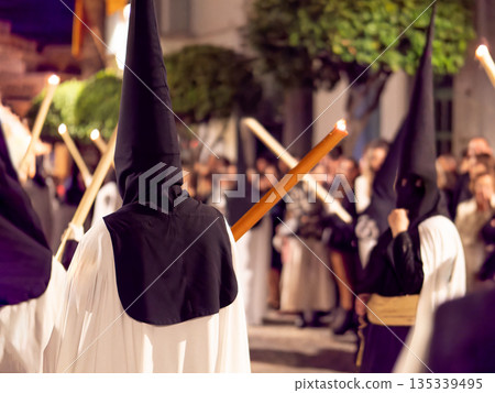 Nazarenes in white tunics and black hoods walking in a night procession with candles. 135339495