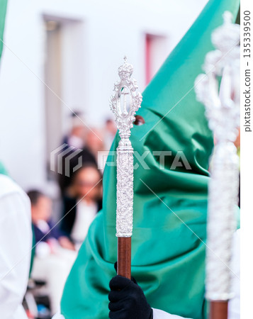 Close up of a nazarene in green hood holding a silver ceremonial procession cross. 135339500