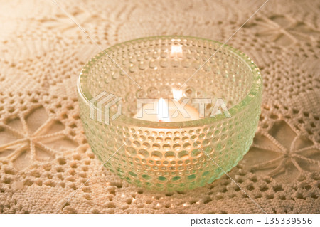 Lit candle in a textured glass holder glowing warmly on a handmade lace tablecloth 135339556