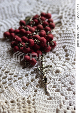Rosary with red wooden beads and a detailed metal crucifix laying on a white crocheted tablecloth Rosary with red wooden beads and a detailed metal crucifix laying on a white crocheted tablecloth 135339634
