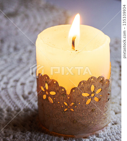 Lit candle glowing on a soft knitted blanket to evoke cozy hygge, relaxation and warm ambiance Lit candle glowing on a soft knitted blanket to evoke cozy hygge, relaxation and warm ambiance 135339934