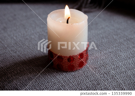 White candle burning with a warm flame, decorated with a red ribbon featuring dark red polka dots 135339990