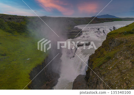 Gullfoss waterfall in Iceland flows in two tiers into a canyon, surrounded by green cliffs. Mist rises as the sky glows with pink and blue hues. 135340100