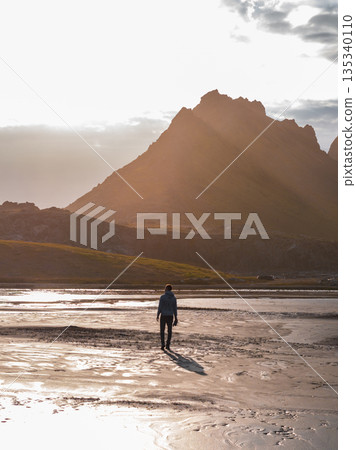 A person walks on wet sand reflecting golden light, with jagged mountains silhouetted in the background near Iceland's Stokknes Peninsula. A person walks on wet sand reflecting golden light, with jagged mountains silhouetted in the background near Iceland's Stokknes Peninsula. 135340110