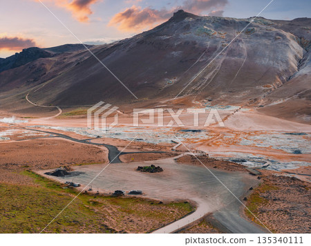 Hverir geothermal area near Lake Myvatn, Iceland, with steaming fumaroles, sulfur streaked earth, a mountain, and a parking area at sunset. 135340111