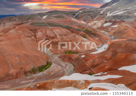 A vibrant aerial view of Iceland's Rainbow Mountains with red, orange, and brown hues, snow patches, a winding river, and a dramatic sunset sky. A vibrant aerial view of Iceland's Rainbow Mountains with red, orange, and brown hues, snow patches, a winding river, and a dramatic sunset sky. 135340113