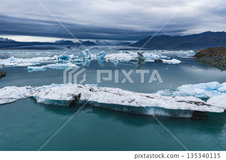 Large icebergs float in turquoise water at Jokulsarlon Glacier Lagoon, framed by mountains and an overcast sky. People stand on the rocky shoreline. Large icebergs float in turquoise water at Jokulsarlon Glacier Lagoon, framed by mountains and an overcast sky. People stand on the rocky shoreline. 135340115