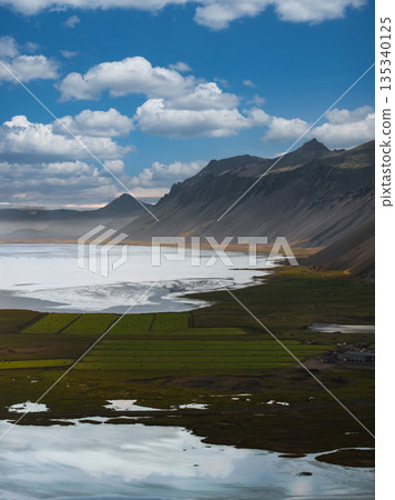 Aerial view of Stokknes Peninsula in Iceland, showing rugged peaks, serene coastline, reflective water, green fields, and a partly cloudy sky. Aerial view of Stokknes Peninsula in Iceland, showing rugged peaks, serene coastline, reflective water, green fields, and a partly cloudy sky. 135340125