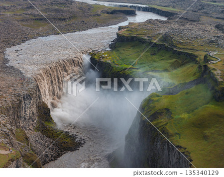 Dettifoss waterfall cascades into a deep canyon, surrounded by cliffs and moss covered terrain. Mist rises prominently, with the river winding upstream. Dettifoss waterfall cascades into a deep canyon, surrounded by cliffs and moss covered terrain. Mist rises prominently, with the river winding upstream. 135340129