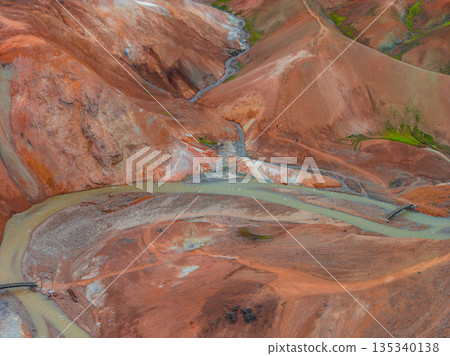Aerial view of Iceland's Rainbow Mountains with vibrant red, orange, and brown hues. A winding river and wooden bridges traverse the rugged terrain. 135340138