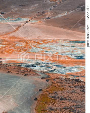 A vibrant geothermal area in Iceland featuring orange and white mineral deposits, steaming vents, and a wooden walkway winding through the barren terrain. A vibrant geothermal area in Iceland featuring orange and white mineral deposits, steaming vents, and a wooden walkway winding through the barren terrain. 135340140