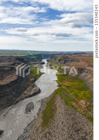 Aerial view of Studlagil Canyon in Iceland, featuring steep basalt columns, a winding silty river, green vegetation, and a partly cloudy sky. Aerial view of Studlagil Canyon in Iceland, featuring steep basalt columns, a winding silty river, green vegetation, and a partly cloudy sky. 135340143