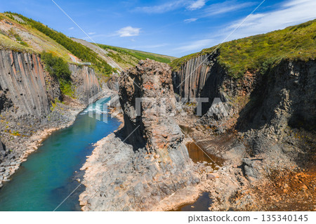 Studlagil Canyon in Iceland features dramatic basalt columns, a vibrant turquoise river, lush green hills, and a clear blue sky in a volcanic landscape. 135340145