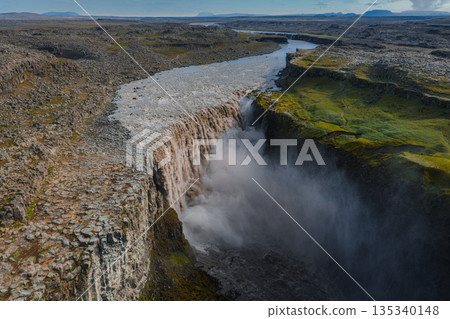 Dettifoss waterfall in Iceland plunges into a deep canyon, surrounded by basalt cliffs, green moss, and mist rising from the powerful flow of water. 135340148