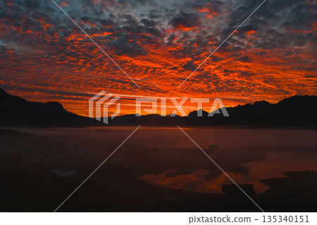 A vibrant red and orange sunset illuminates the sky over silhouetted Icelandic peaks. Mist blankets the landscape, with reflections in a tranquil water surface. 135340151