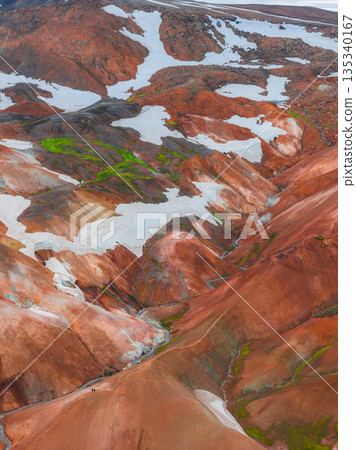 Vibrant rhyolite mountains in Landmannalaugar, Iceland, with red, orange, and brown hues, snow patches, green moss, and two small figures visible. Vibrant rhyolite mountains in Landmannalaugar, Iceland, with red, orange, and brown hues, snow patches, green moss, and two small figures visible. 135340167