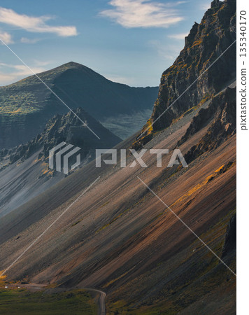 A dramatic Icelandic mountainside with steep ridges, rocky and grassy textures, a winding road below, and peaks under a clear sky with scattered clouds. A dramatic Icelandic mountainside with steep ridges, rocky and grassy textures, a winding road below, and peaks under a clear sky with scattered clouds. 135340170