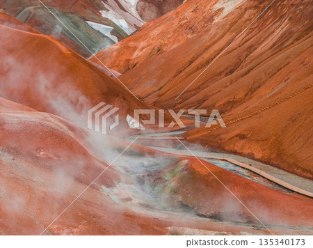 Vibrant red and orange rhyolite mountains with steam rising from geothermal vents, a wooden pathway, and snow patches in Kerlingarfjoll, Iceland. 135340173