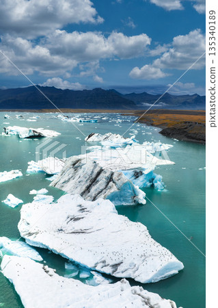 Aerial view of Jokulsarlon Glacier Lagoon in Iceland, featuring white icebergs in turquoise water, rugged mountains, and a clear blue sky with clouds. Aerial view of Jokulsarlon Glacier Lagoon in Iceland, featuring white icebergs in turquoise water, rugged mountains, and a clear blue sky with clouds. 135340189