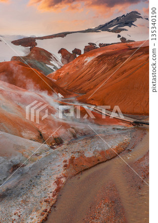 Kerlingarfjoll's red and orange rhyolite mountains with steam vents, a wooden bridge over a stream, snow capped peaks, and a sunset glow. Kerlingarfjoll's red and orange rhyolite mountains with steam vents, a wooden bridge over a stream, snow capped peaks, and a sunset glow. 135340190