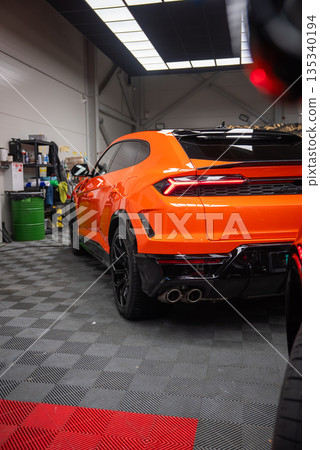 A three quarter rear view of an orange Lamborghini Urus SE parked indoors, showing sharp taillights, quad exhaust tips, black diffuser, and gloss black wheels. 135340194