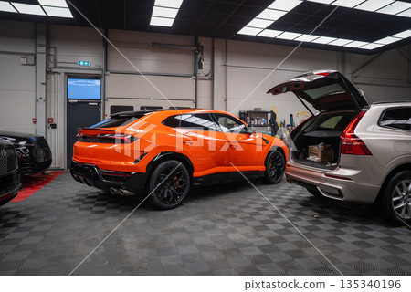 An orange Lamborghini Urus SE stands on a gray checkered floor, under bright panel lighting, with detailing tools along the wall and nearby cars including a silver SUV. 135340196