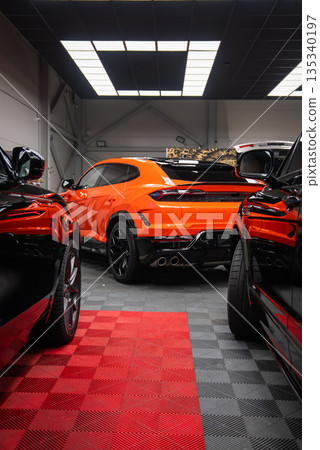 A bright orange Lamborghini Urus SE is parked indoors between dark SUVs. Quad exhausts, black diffuser, and sharp taillights stand out under industrial ceiling lights. 135340197