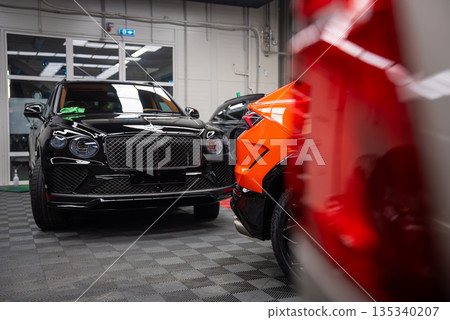 Close up indoor workshop scene shows orange Lamborghini Urus SE parked by a glossy black Bentley Bentayga, with a red car blurred in front, under bright overhead lights. 135340207