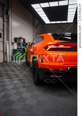Orange Lamborghini Urus SE in a detailing bay, low rear three quarter view shows quad exhausts, angular taillights, gloss black diffuser, panel lighting, and checkered floor. 135340224