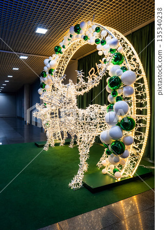 Two illuminated reindeer leap through a light wrapped arch in Riga, Latvia, on green turf with reflective tile flooring and a gridded ceiling, shot from a low angle. 135340238