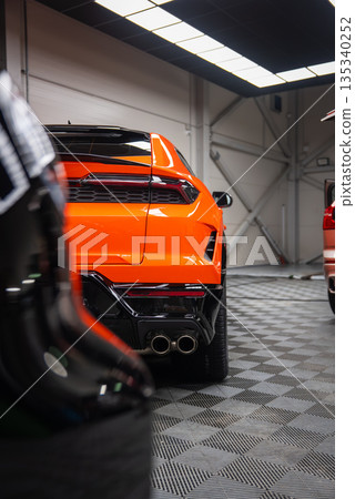 Vivid orange Lamborghini Urus SE rear quarter in a workshop, quad exhausts, sharp taillights, glossy black diffuser, checkered rubber floor, other cars in black and red 135340252