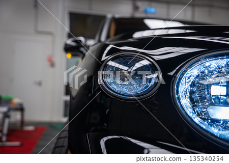 Close up shows front left headlight cluster of a black Bentley Bentayga Speed indoors, with twin circular LED lamps and crystalline detailing reflecting overhead lights. 135340254