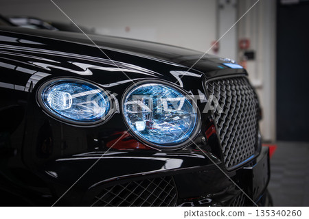 Close view of a black Bentley Bentayga Speed in a garage, twin crystal cut LED headlights with blue tinted illumination and diamond mesh grille with winged badge. Close view of a black Bentley Bentayga Speed in a garage, twin crystal cut LED headlights with blue tinted illumination and diamond mesh grille with winged badge. 135340260