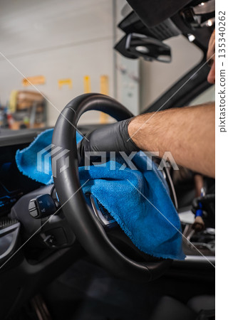 A person in black nitrile gloves wipes a car steering wheel with a bright blue microfiber cloth inside an auto detailing shop, soft indoor light and shallow depth of field A person in black nitrile gloves wipes a car steering wheel with a bright blue microfiber cloth inside an auto detailing shop, soft indoor light and shallow depth of field 135340262