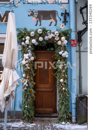 A wooden doorway with evergreen garlands, white baubles, and gold accents sits beneath a cow mural in Old Riga, Latvia, with light snow and a wrought iron lantern. A wooden doorway with evergreen garlands, white baubles, and gold accents sits beneath a cow mural in Old Riga, Latvia, with light snow and a wrought iron lantern. 135340264