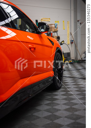 Bright orange Lamborghini Urus SE sits in a workshop, glossy paint reflects ceiling lights, black alloy wheel and side skirt in frame, tools and gear line gray wall. 135340273