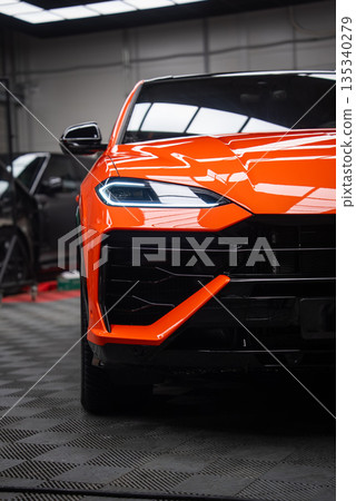 Close up shows orange Lamborghini Urus SE in an indoor workshop, with angular LED headlight, sculpted hood, black grille and orange aero blade under bright lights. 135340279