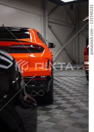 Orange Lamborghini Urus SE sits indoors on gray checkered floor, rear three quarter view shows quad exhausts, sharp tail lights, sculpted bumper, cars parked alongside. 135340281