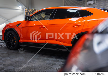 A close up side view shows an orange Lamborghini Urus SE parked in a modern workshop, with glossy paint, black wheels, orange calipers, checkered flooring, and detailing gear. 135340282