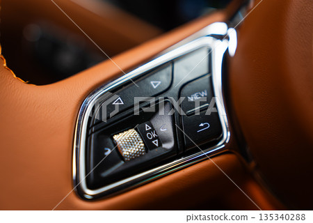 Right side controls of a Bentley Bentayga Speed show a knurled roller, arrow buttons, and a VIEW key, with tan leather and chrome trim in warm directional light. 135340288
