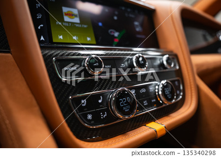 Close up interior view shows carbon fiber trim, knurled dials, tan leather, and contrast stitching. Screen shows radio and navigation. Climate reads 22 C with seat heating. Close up interior view shows carbon fiber trim, knurled dials, tan leather, and contrast stitching. Screen shows radio and navigation. Climate reads 22 C with seat heating. 135340298