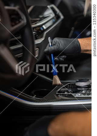 A gloved hand uses a detailing brush to clean dust around the gear selector and control dials on carbon fiber trim, inside a modern car with soft focused lighting. A gloved hand uses a detailing brush to clean dust around the gear selector and control dials on carbon fiber trim, inside a modern car with soft focused lighting. 135340300