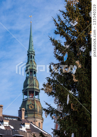 A Christmas tree with geometric ornaments stands before Riga Cathedral in Old Town Riga, Latvia. Daylight winter scene shows snow on roofs and the gilded rooster. A Christmas tree with geometric ornaments stands before Riga Cathedral in Old Town Riga, Latvia. Daylight winter scene shows snow on roofs and the gilded rooster. 135340319