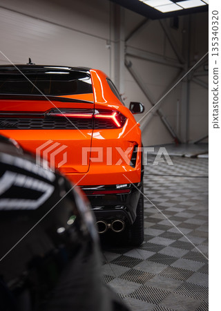 Three quarter rear view shows orange Lamborghini Urus SE in a workshop with angular LED taillight, quad exhaust tips, checkered floor, and overhead fluorescent lights. 135340320