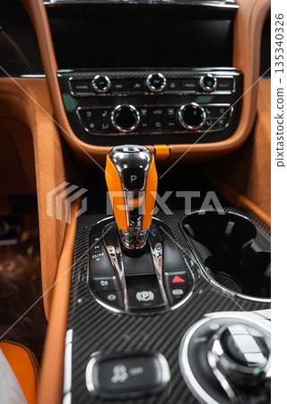 Close up indoor view of a Bentley Bentayga Speed center console, crystal and metal gear selector with P indicator, carbon fiber trim, tan leather, knobs, switches, cup holders. Close up indoor view of a Bentley Bentayga Speed center console, crystal and metal gear selector with P indicator, carbon fiber trim, tan leather, knobs, switches, cup holders. 135340326